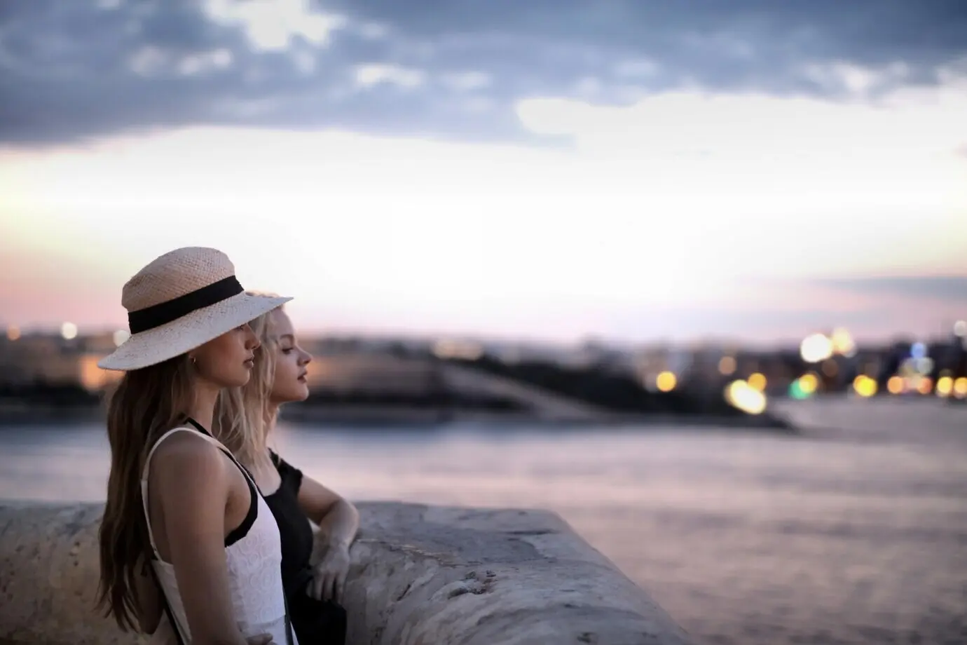 Two young females stand beside the stone border, gazing at the blurry harbor.