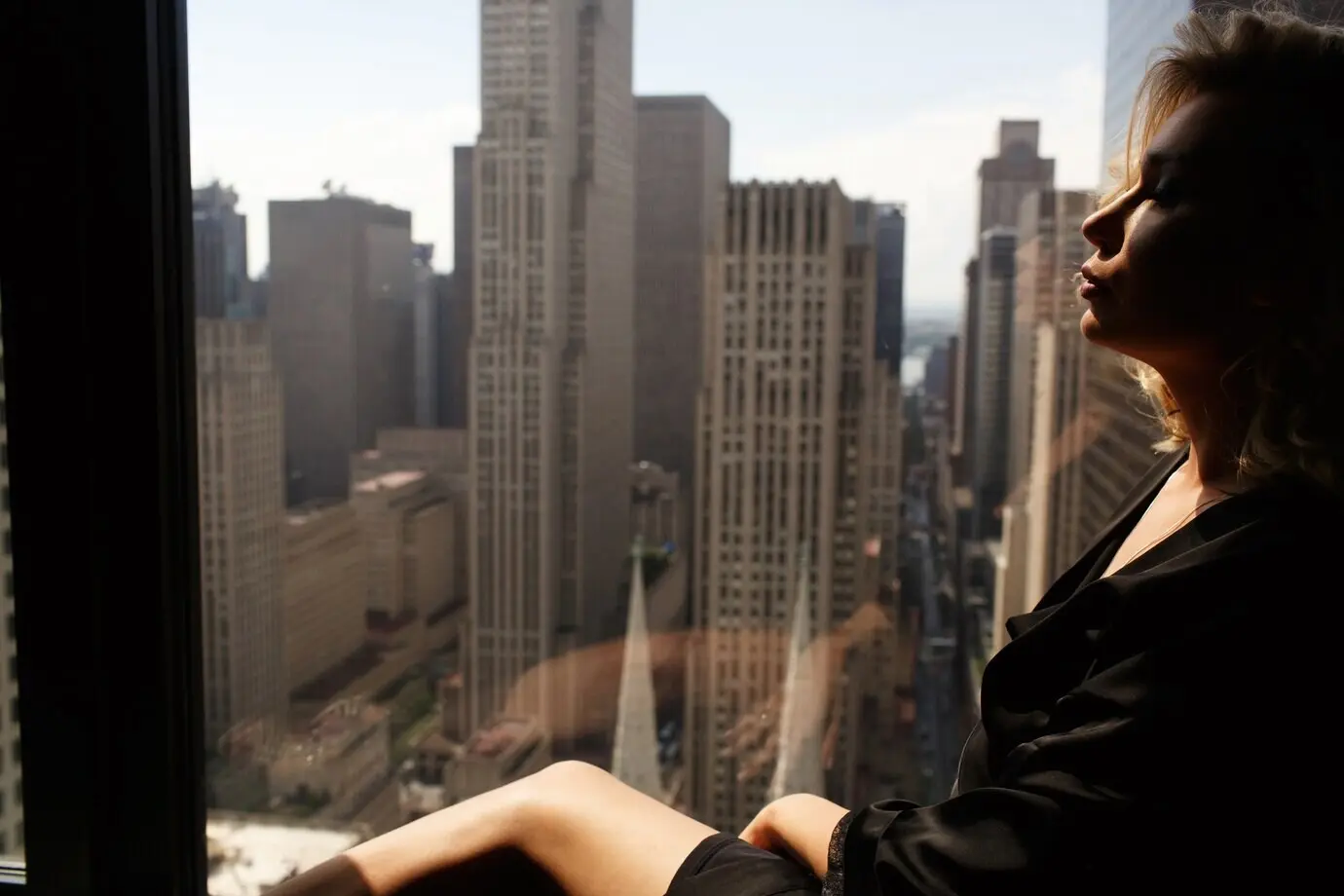 A pensive woman in a black silk robe sits on a windowsill in front of a beautiful New York cityscape.